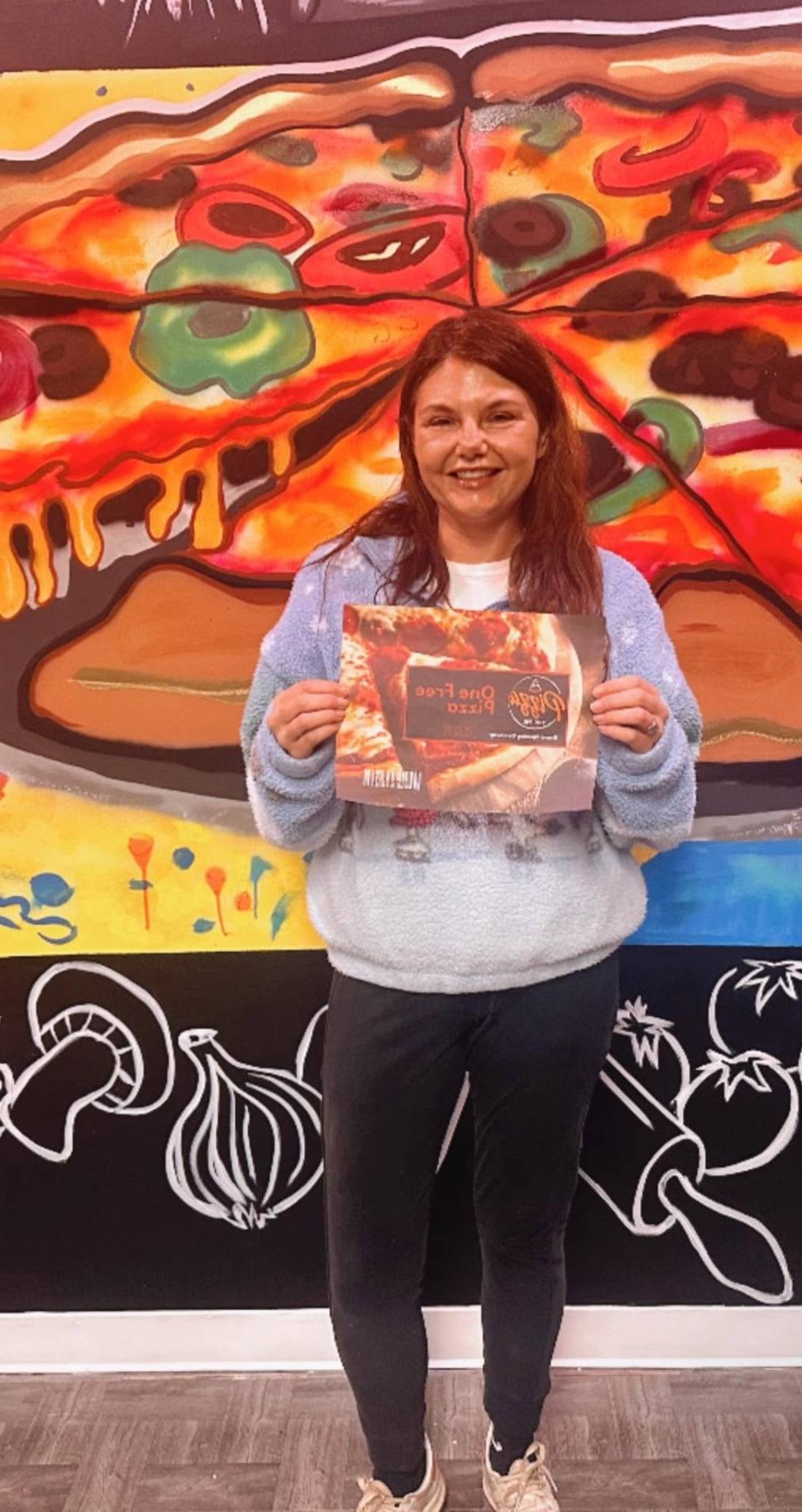 Guest holding a free pizza card in front of the mural at The Spot on 10th